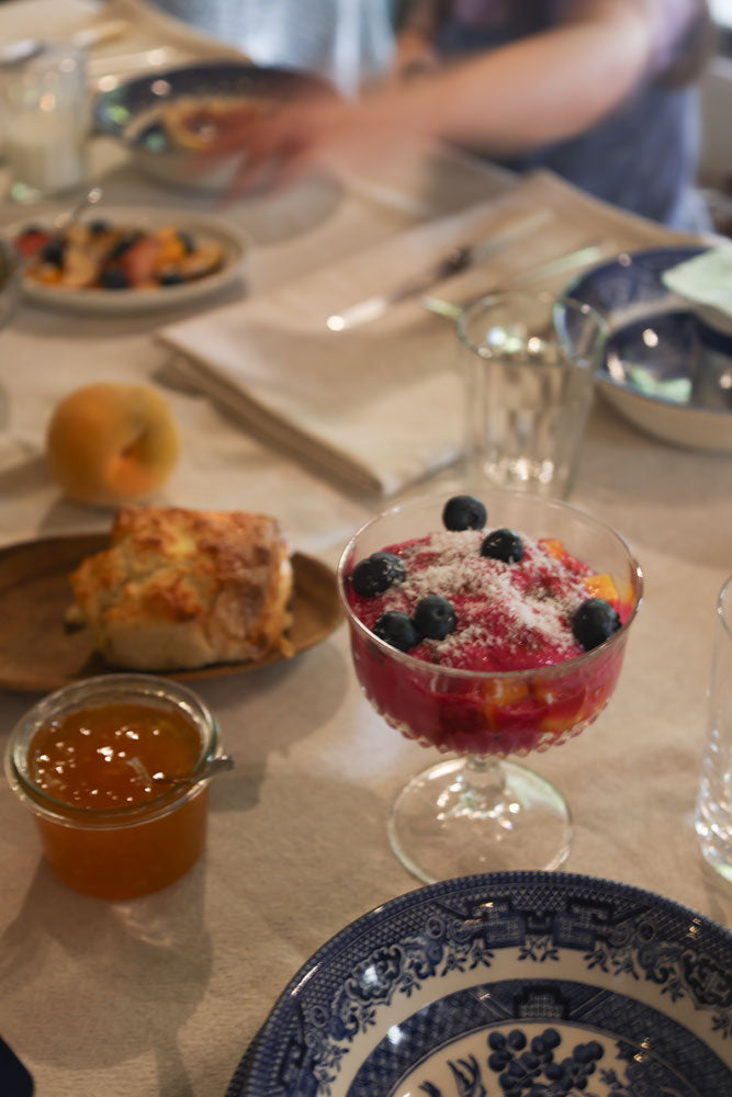 A dessert glass filled with red fruit, blueberries, and white sugar on a dining table, alongside a small jar of orange jam, a pastry, and other homestead breakfast items at San Gabriel.